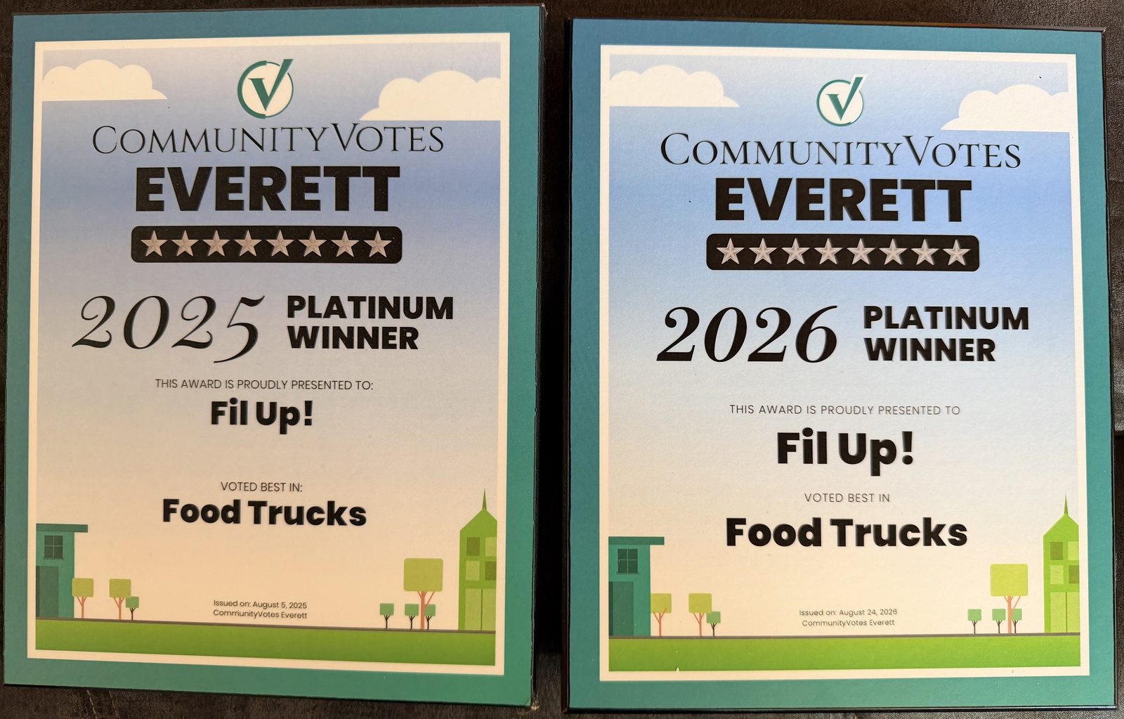 Two CommunityVotes Everett award plaques lying side by side on a dark leather seat. The left one reads 2025 Platinum Winner and the right one 2026 Platinum Winner; both are presented to Fil Up! and both read “Voted best in: Food Trucks”
