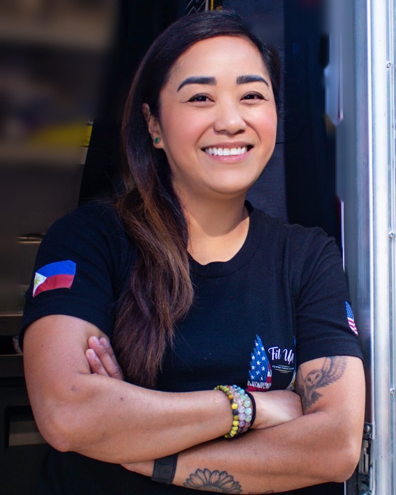 Smiling at the camera with her arms folded in the doorway of the truck, in a black Fil Up! shirt with a Philippine flag patch on the sleeve