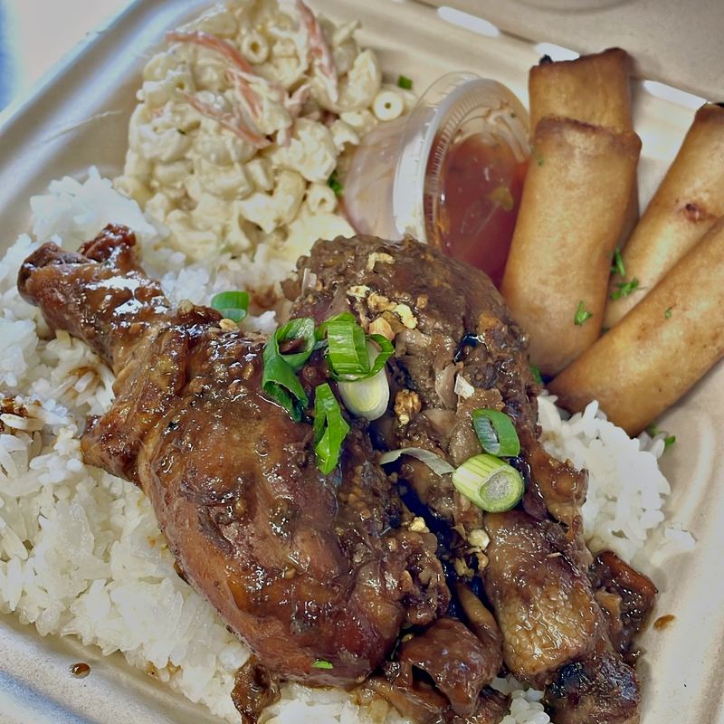 A takeout box with 3 partitions: the big compartment in the bottom-middle contains 2 chicken legs braised with dark glossy sauce and scattered green onions on top, all sitting on steamed white rice; the smaller, top-right compartment contains 4 tightly rolled, golden brown lumpia fresh from the fryer, with a small, closed cup of housemade sweet chili dipping sauce on the side; the smaller, top-left compartment contains macaroni salad.