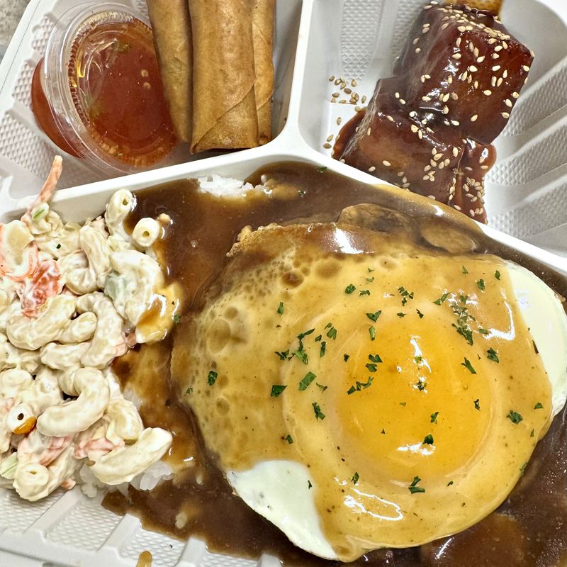 A takeout box with 3 partitions: the big compartment in the bottom-middle contains a runny-yolked fried egg covered in brown gravy and scattered herbs on top of a beef patty sitting on steamed white rice, with macaroni salad on the side; the smaller, top-left compartment contains 3 tightly rolled, golden brown lumpia fresh from the fryer, with a small, closed cup of housemade sweet chili dipping sauce on the side; the smaller, top-right compartment contains 2 thick-cut, dark glazed, sesame-flecked pork belly chunks.