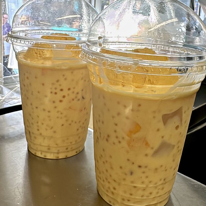 Two clear domed-lid cups, beaded with condensation, holding a pale gold creamy mango base with small sago pearls and pale mango chunks spread throughout, and deeper-gold mango ice cream resting on top of the base under the lid.