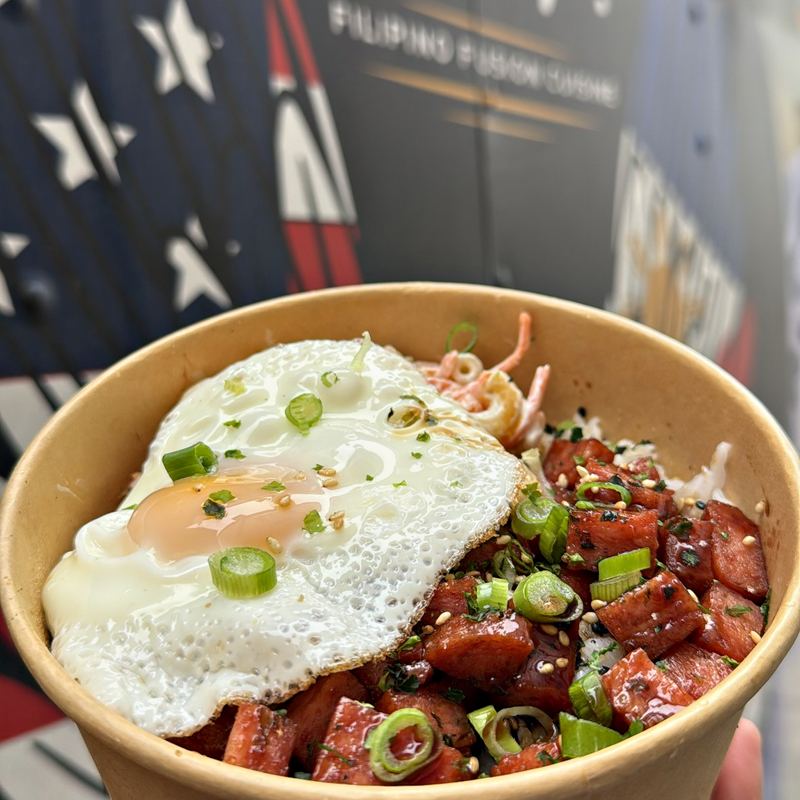 A runny-yolked fried egg sitting atop diced Spam seared dark at the edges over a bowl of steamed white rice with green onions and sesame scattered across, held up outside the food truck trailer.