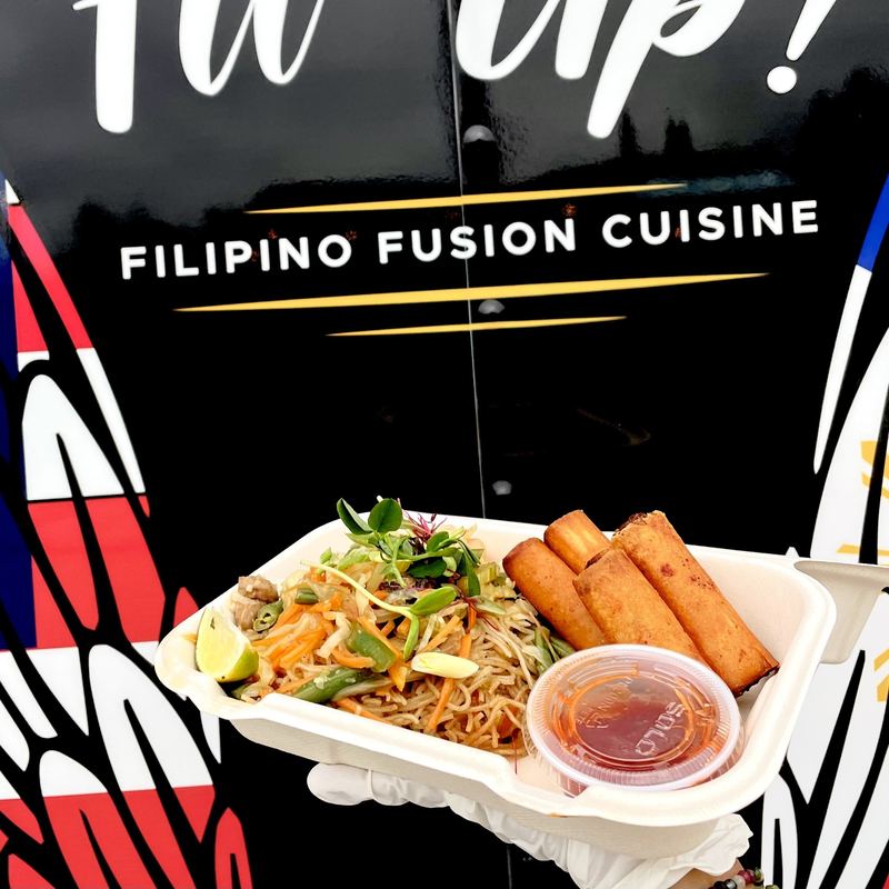 Pancit noodles coiled in a takeout box under a tuft of vegetables and greens, with 4 tightly rolled, golden brown lumpia fresh from the fryer, with a small, closed cup of housemade sweet chili dipping sauce on the side, held up outside the food truck trailer.