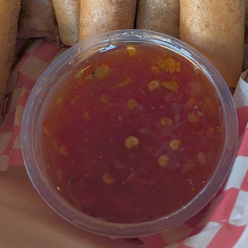 A clear cup of deep red sweet chili sauce in a basket of lumpia on red-checkered paper. Chili seeds and flecks of garlic are suspended through it.
