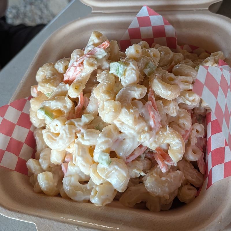 A heaped scoop of creamy macaroni salad flecked with carrots, in a takeout box lined with red-checkered paper.