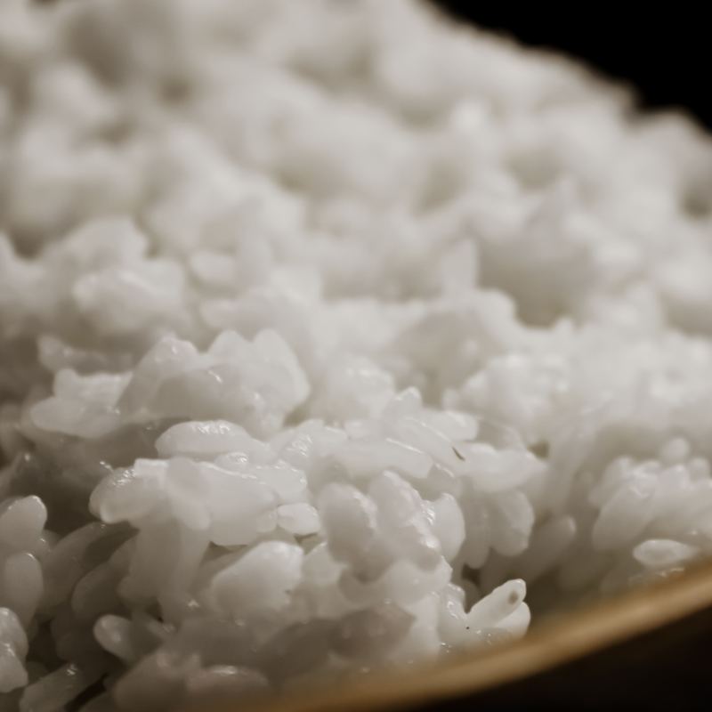 Steamed white rice in close-up with every grain separate and glossy. The polished rim of a dark wooden bowl curves across the lower right corner.