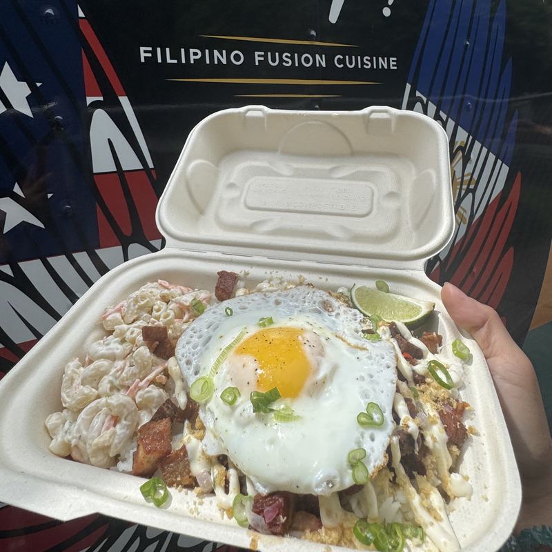 A hand holds an open takeout box in front of the truck, below lettering on the wrap that reads Filipino Fusion Cuisine. Inside the box, a sunny-side-up egg sits over crisped sisig pork belly and steamed rice, drizzled with cilantro-calamansi aioli, scattered with sliced green onion, and banked with macaroni salad, with a lime wedge at the near corner.