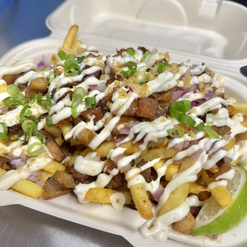 Golden French fries buried under chopped crisped sisig pork belly and red onion, laced with cilantro-calamansi aioli, topped with scattered green onion, and a lime wedge at the edge of the box.