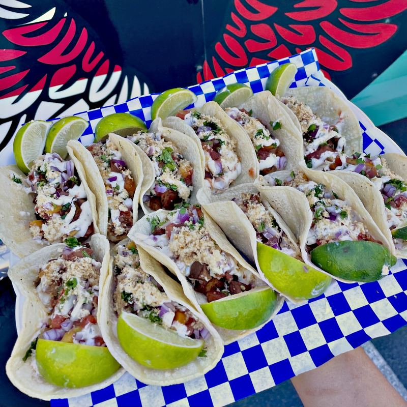 Three sisig tacos in corn tortillas with peach-mango salsa, red onion, crushed chicharrones, and cilantro-calamansi aioli, with lime wedges on the side.