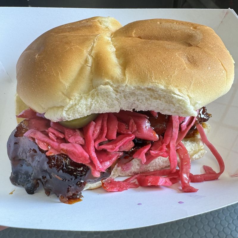 A slider in a soft bun, dark-glazed pork belly spilling out under a tangle of bright purplish-pink pickled cabbage with a jalapeño peeking through.