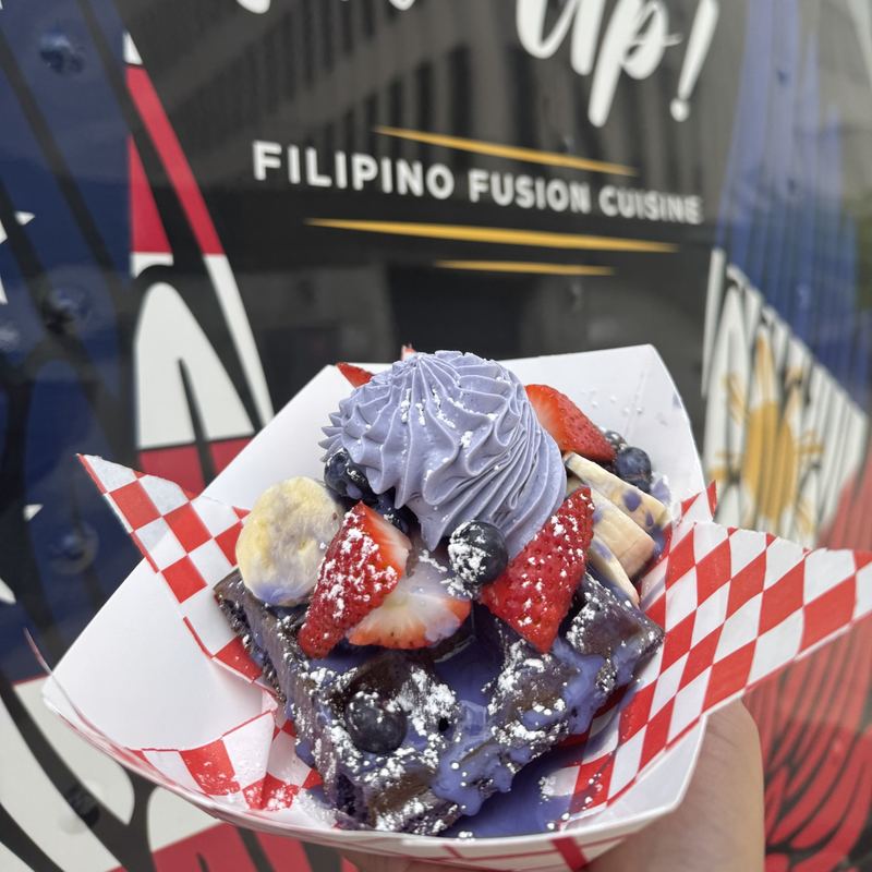 Dark purple ube waffle dusted white with powdered sugar, drizzled with purple ube sweet cream, all under a tall swirl of purple ube whipped cream and piled with cut strawberries, whole blueberries, and sliced bananas.