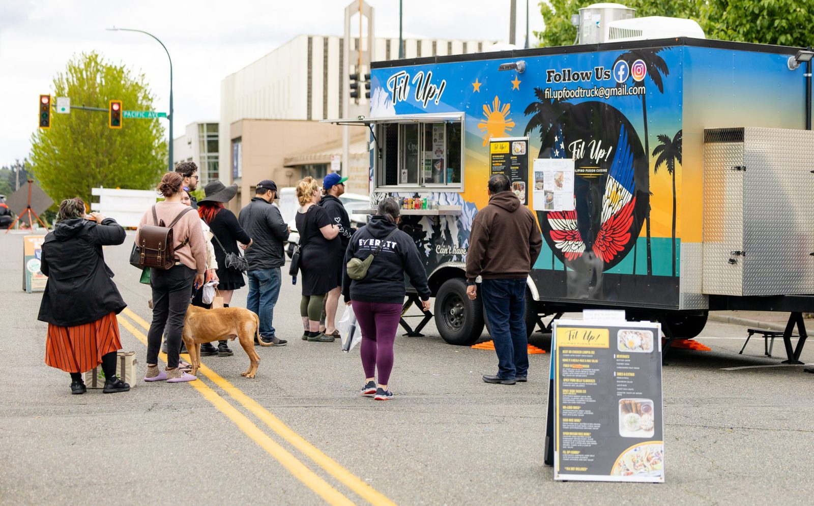 A line of customers waits with their backs to the camera on a closed street, facing the open serving window of the Fil Up! trailer. The trailer wrap carries the Fil Up! name, a gold sunburst, palm silhouettes, and a circle of red, blue, and white wings. An A-frame menu board stands at the right of the frame, and a dog on a leash is partway down the line.