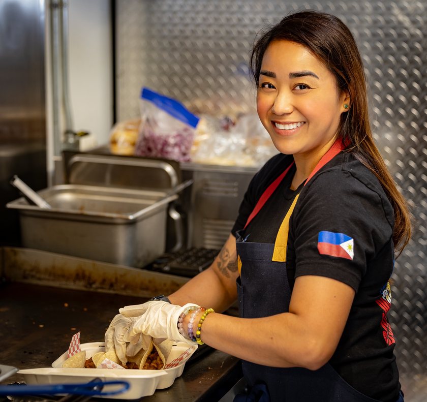 Chef Sarah inside the trailer, smiling toward the camera as she settles a taco into a white takeout box with a gloved hand. Three corn tortillas are filled with reddish-brown sisig pork and separated by red-and-white checkered paper, on a flat-top griddle. She wears a black tee with a Philippine flag patch on the sleeve, a navy apron with a yellow bib strap, and beaded bracelets at her wrist. Behind her are stainless steam-table pans and clear bags of prepped ingredients, one of them sliced red onion, against the trailer’s diamond-plate wall.