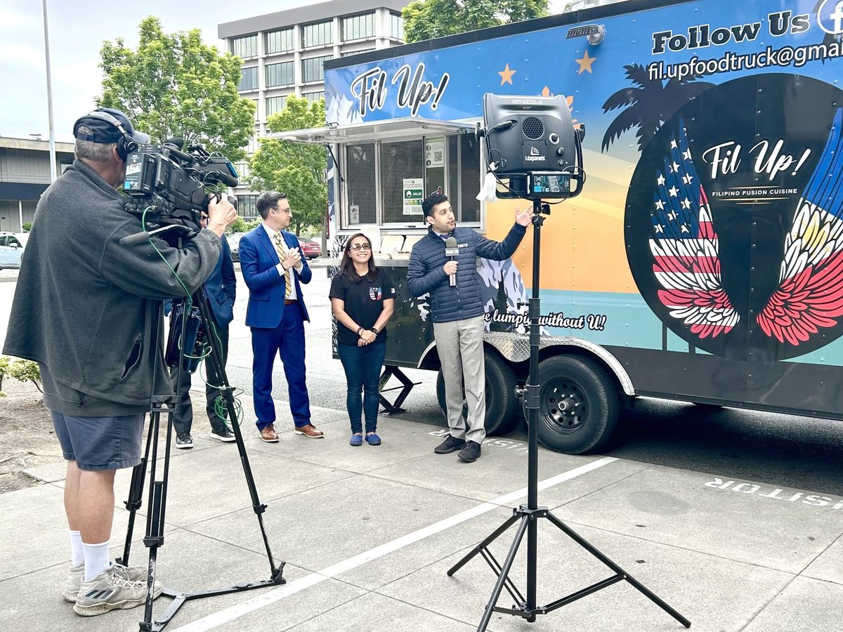 A FOX 13 Seattle reporter speaking to camera beside the Fil Up! trailer while a camera operator films him. Chef Sarah stands between the reporter and a second FOX 13 Seattle reporter, with the serving window open behind them