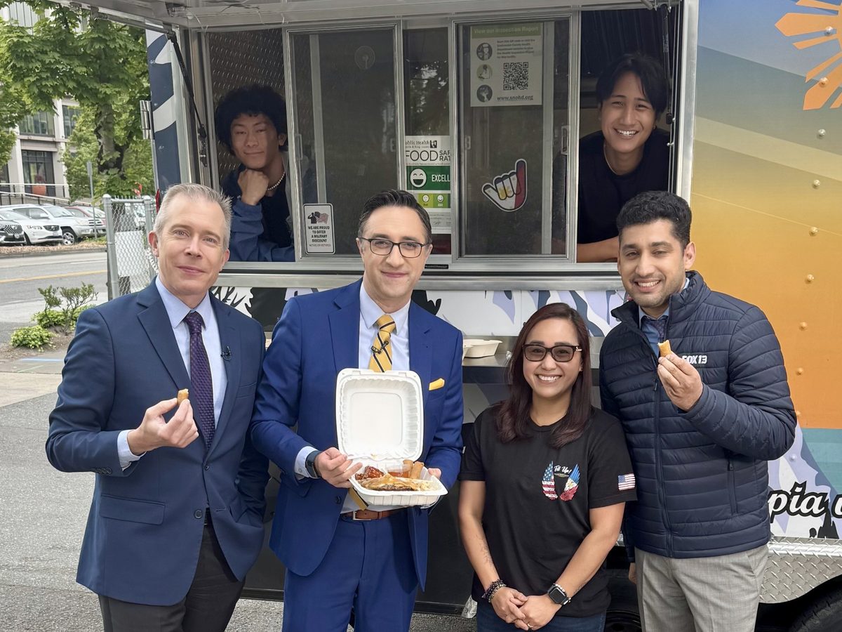 Three FOX 13 Seattle reporters standing with Chef Sarah in front of the Fil Up! trailer. Two of the reporters hold a piece of lumpia; the third holds an open container of food. Behind them Rylan leans out of the left serving window, Sean out of the right