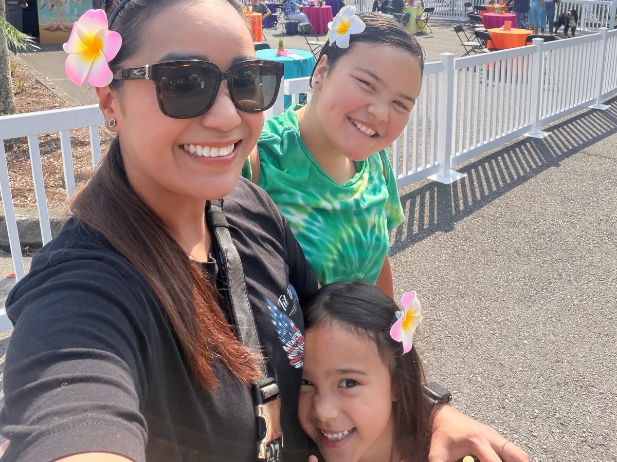 Chef Sarah with her two daughters at the Lynnwood Luau, all three wearing plumeria flowers. Market tents and tables stand behind them