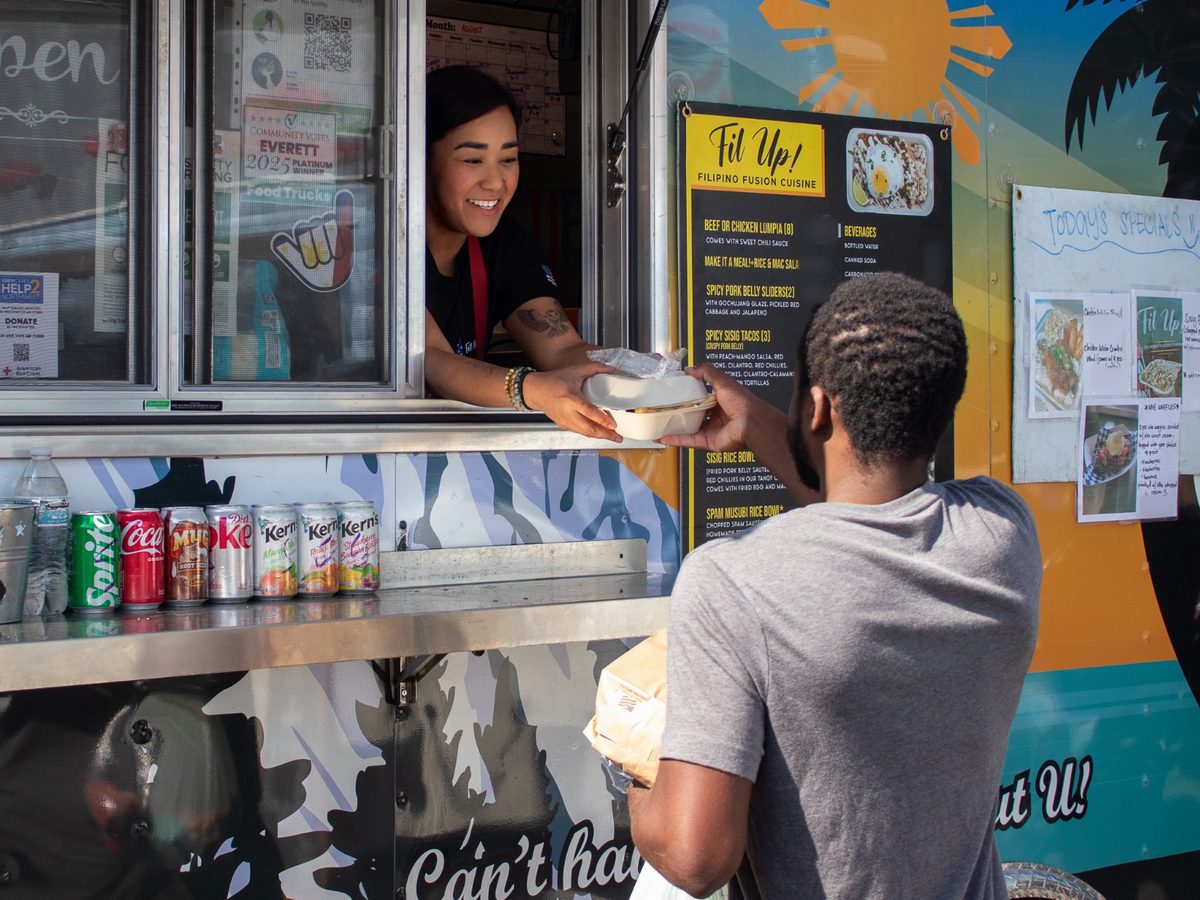 Chef Sarah leaning out of the Fil Up! window, smiling, as she hands a container of food across the counter to a customer