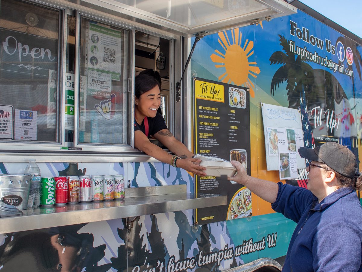 Chef Sarah passing a food container through the window to a customer. The menu board and the Follow Us panel are on the side of the truck beside her